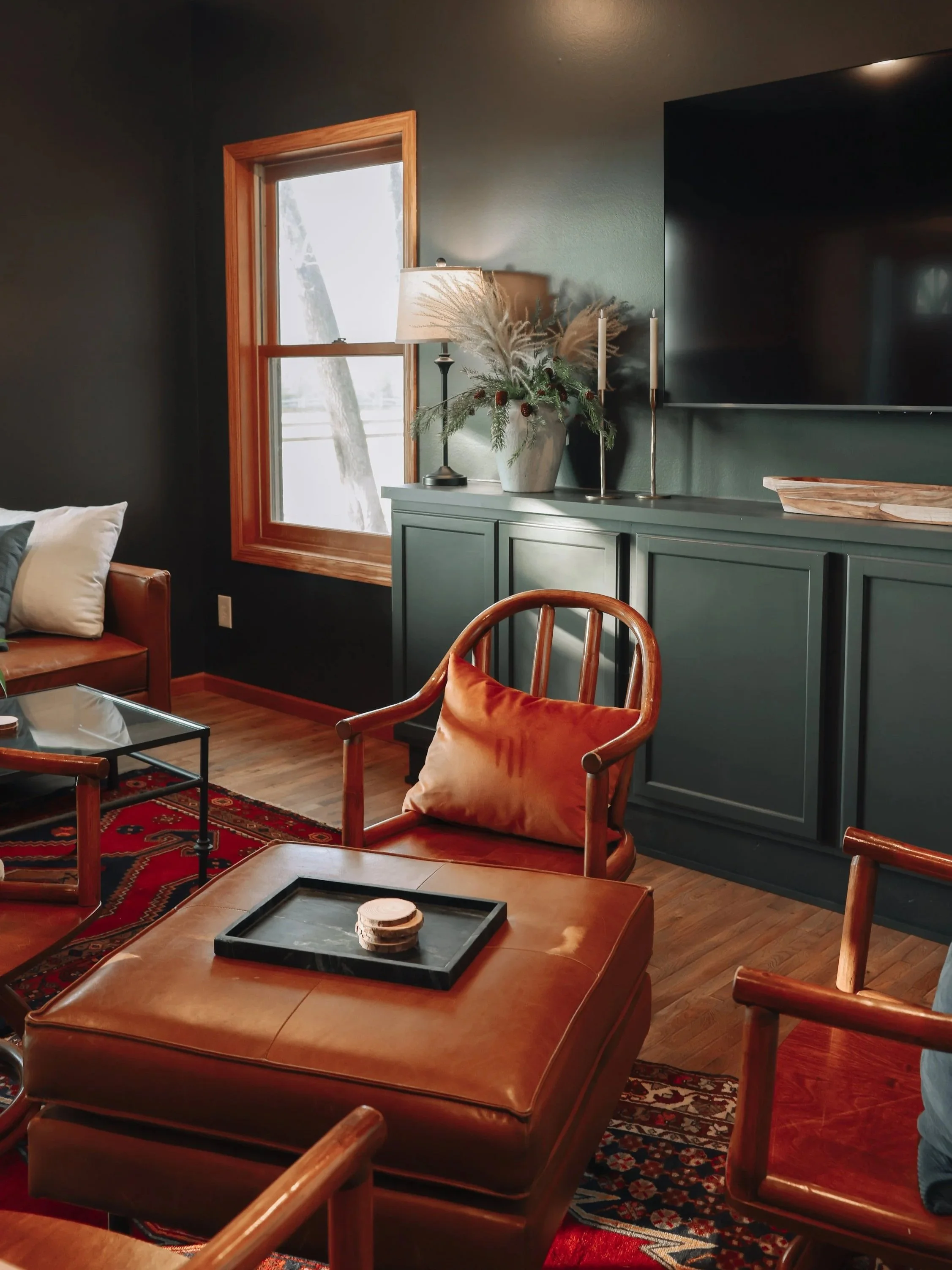 A cozy living room with dark green walls, a wooden-framed window, a teal cabinet, a flat-screen TV, a decorative lamp, and various chairs with cushions, set on a wooden floor and a patterned rug.