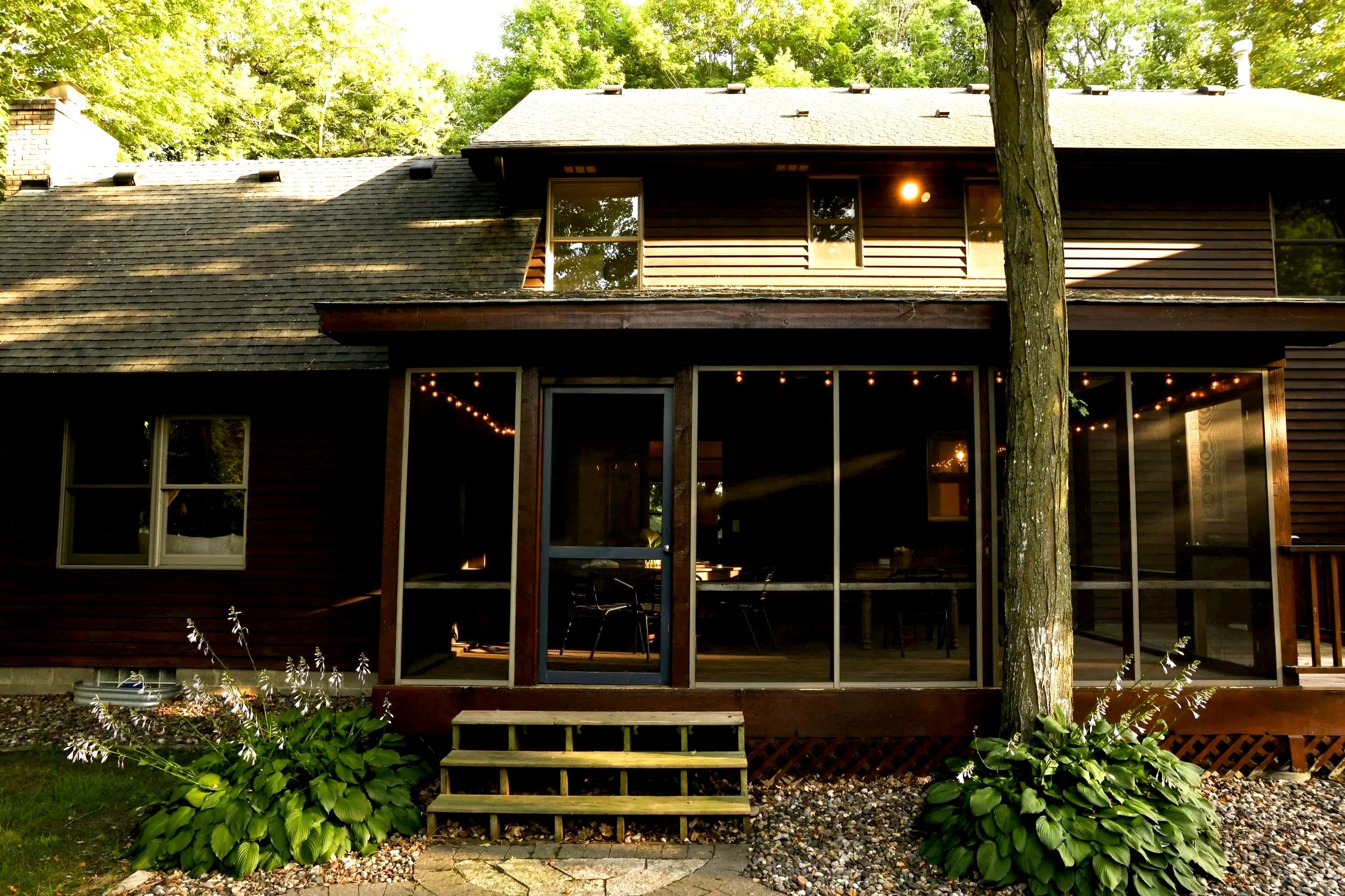 A two-story house with a wooden exterior, large glass sliding doors, and windows, surrounded by green trees and plants. There are steps leading to the porch, and string lights are visible inside through the glass doors.