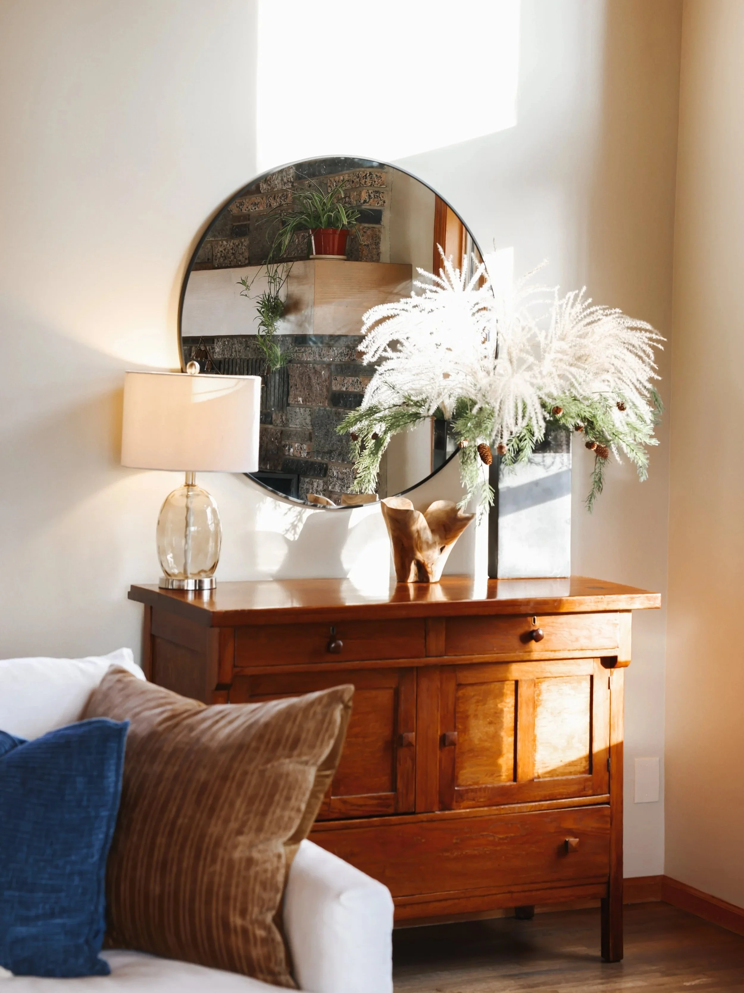 Wooden dresser with a round mirror above it, decorated with a vase of white flowers and pinecones. A lamp with a clear glass base is on the left side of the dresser. The room has white walls and a window with sunlight coming in.