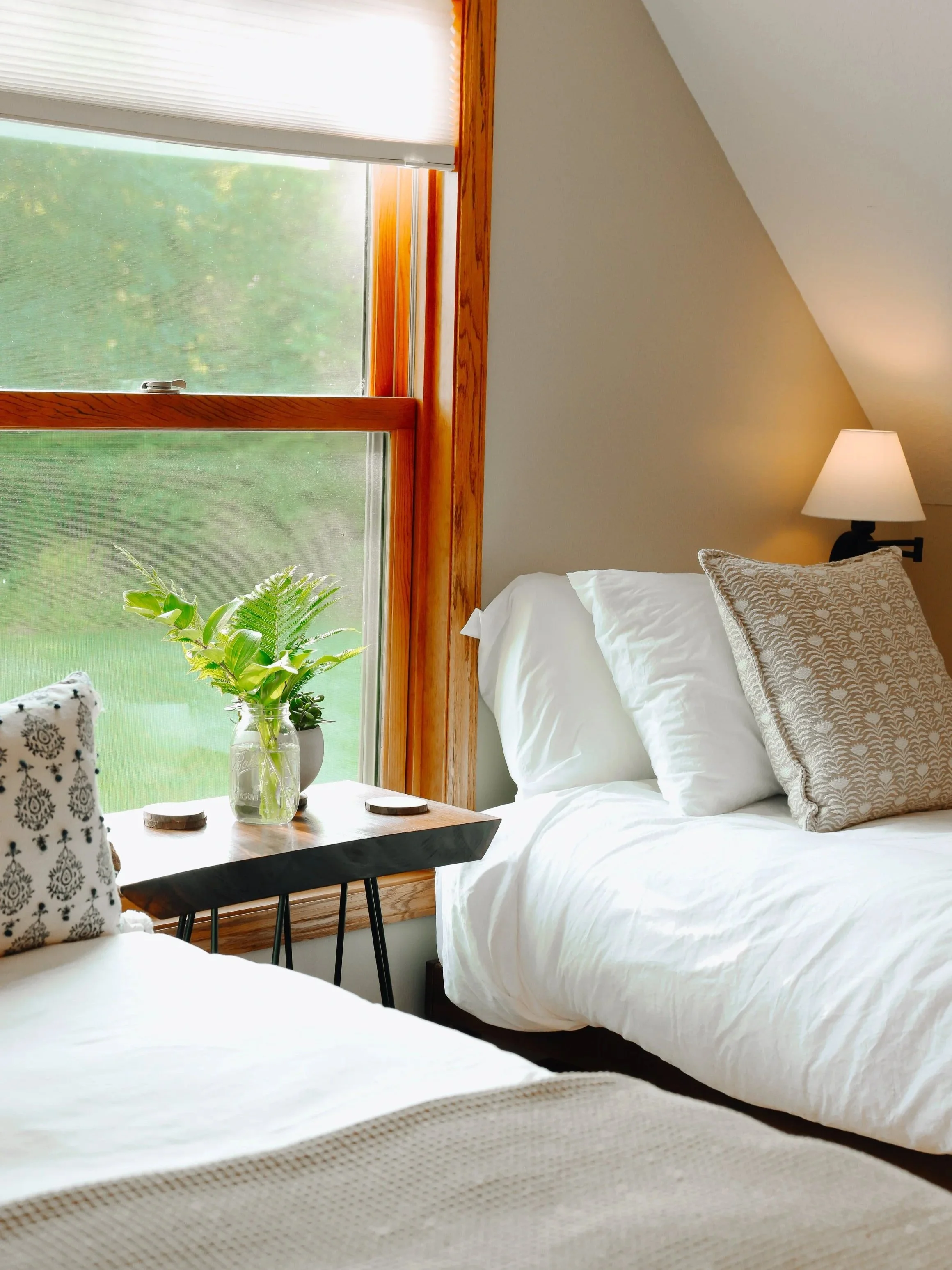 A cozy bedroom with a large window, white bedding, a patterned pillow, a small black bedside table with a glass vase and green plant, and a lamp on the wall.