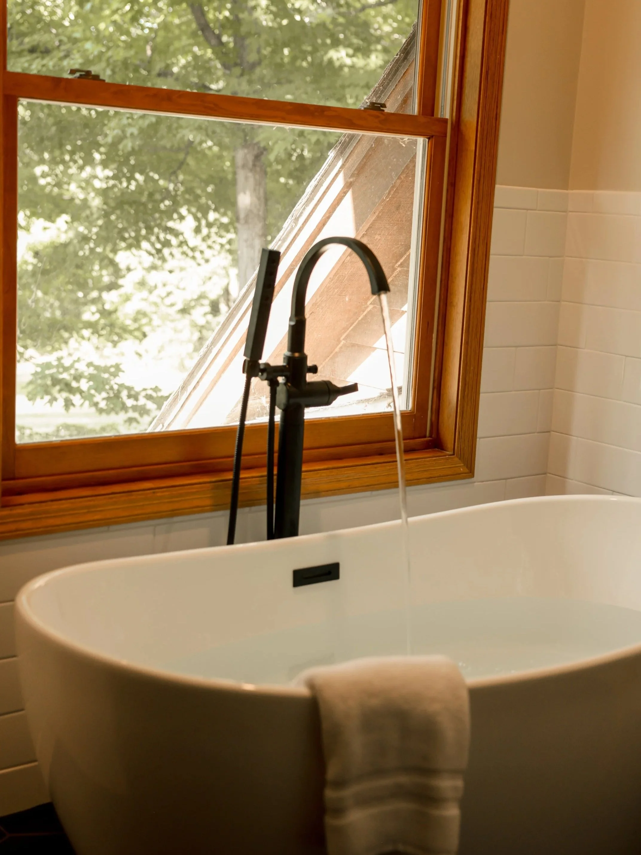 A bathtub with a black faucet sitting in front of a large window with a wooden frame, overlooking green trees outside.