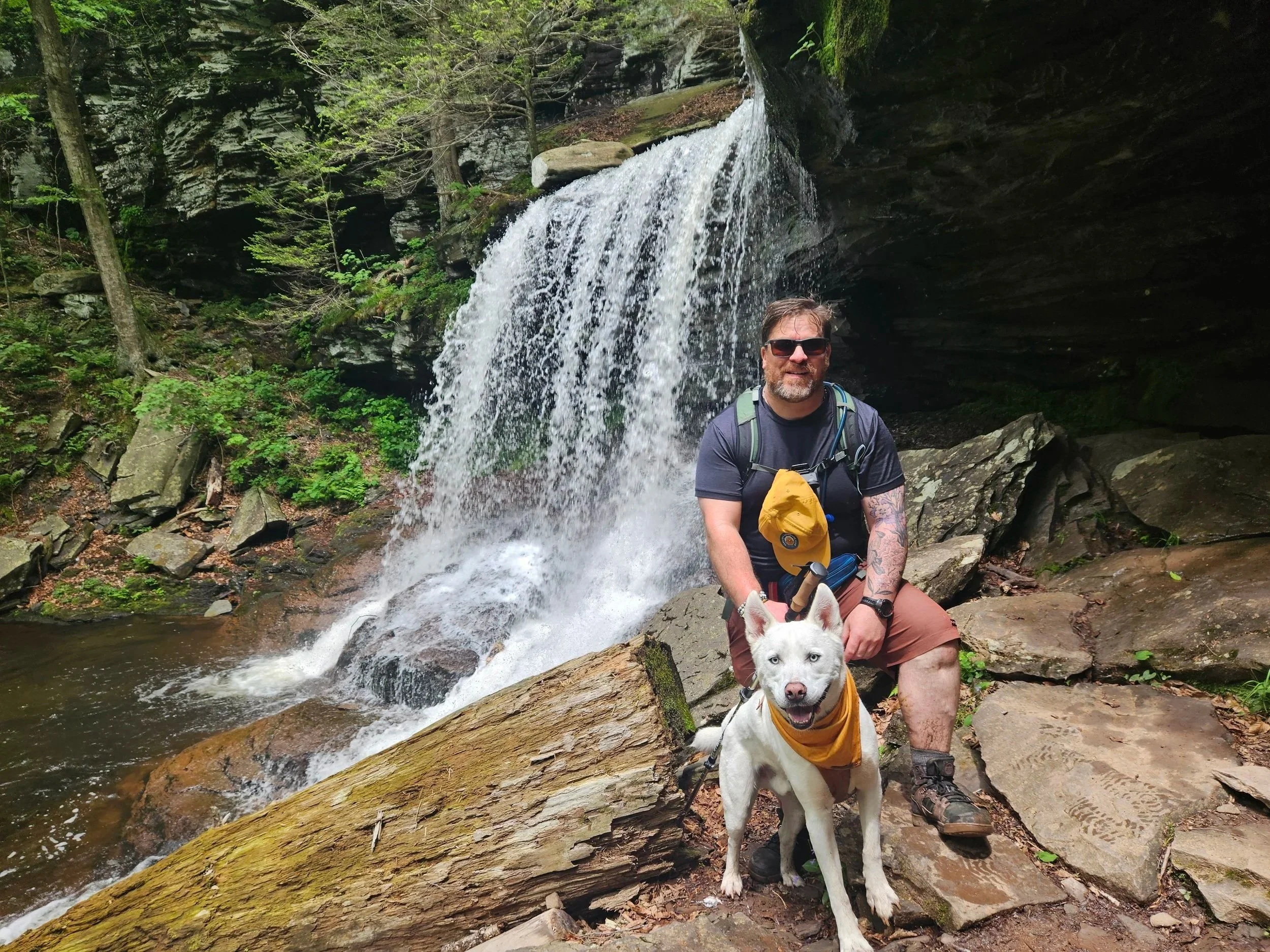A man wearing sunglasses, a backpack, and outdoor clothing is sitting on rocks near a waterfall in a forest. A white dog with a yellow bandana is in front of him, also sitting on rocks, with the waterfall in the background.