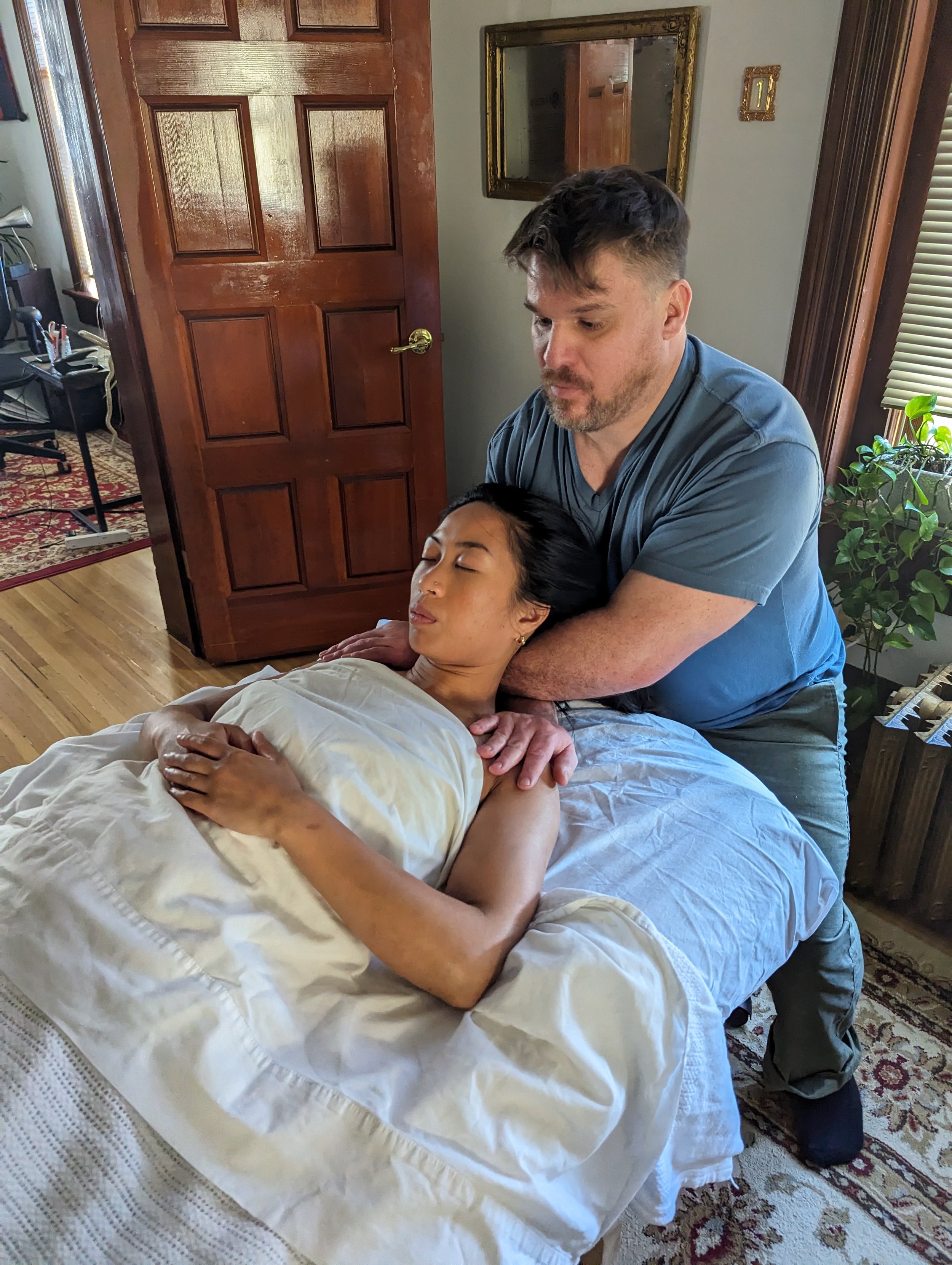 A man giving a woman a massage on a bed in a cozy, well-lit room with wooden furniture and a patterned rug.