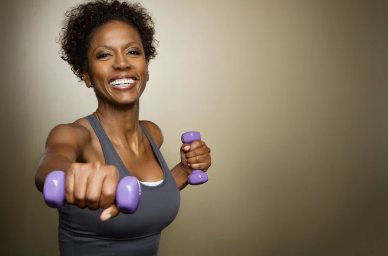 Smiling woman in workout clothes holding purple dumbbells at shoulder height in a fitness studio.