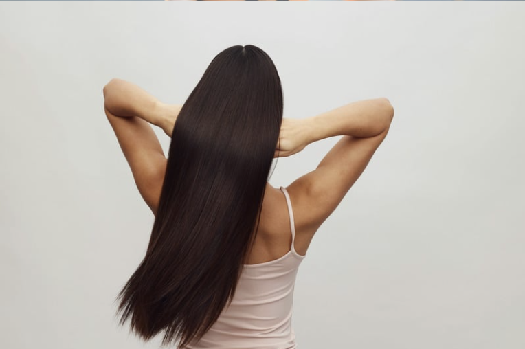 A woman with long, straight, dark hair seen from the back, with her arms raised and hands resting on her head, standing against a plain light gray background.