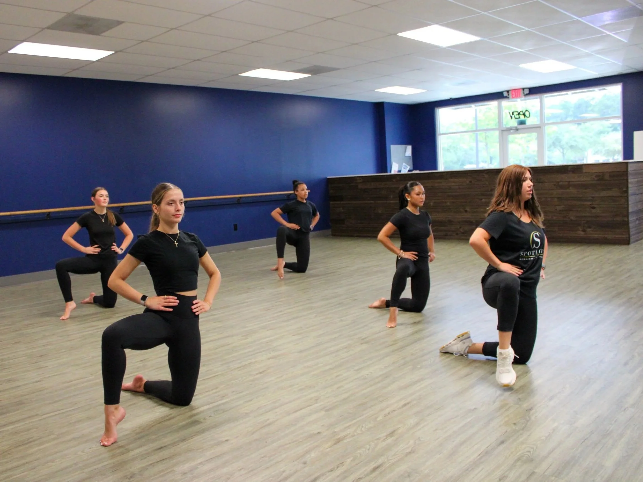 A group of five women in black outfits practicing yoga in a spacious studio with blue walls, wooden flooring, and large windows.