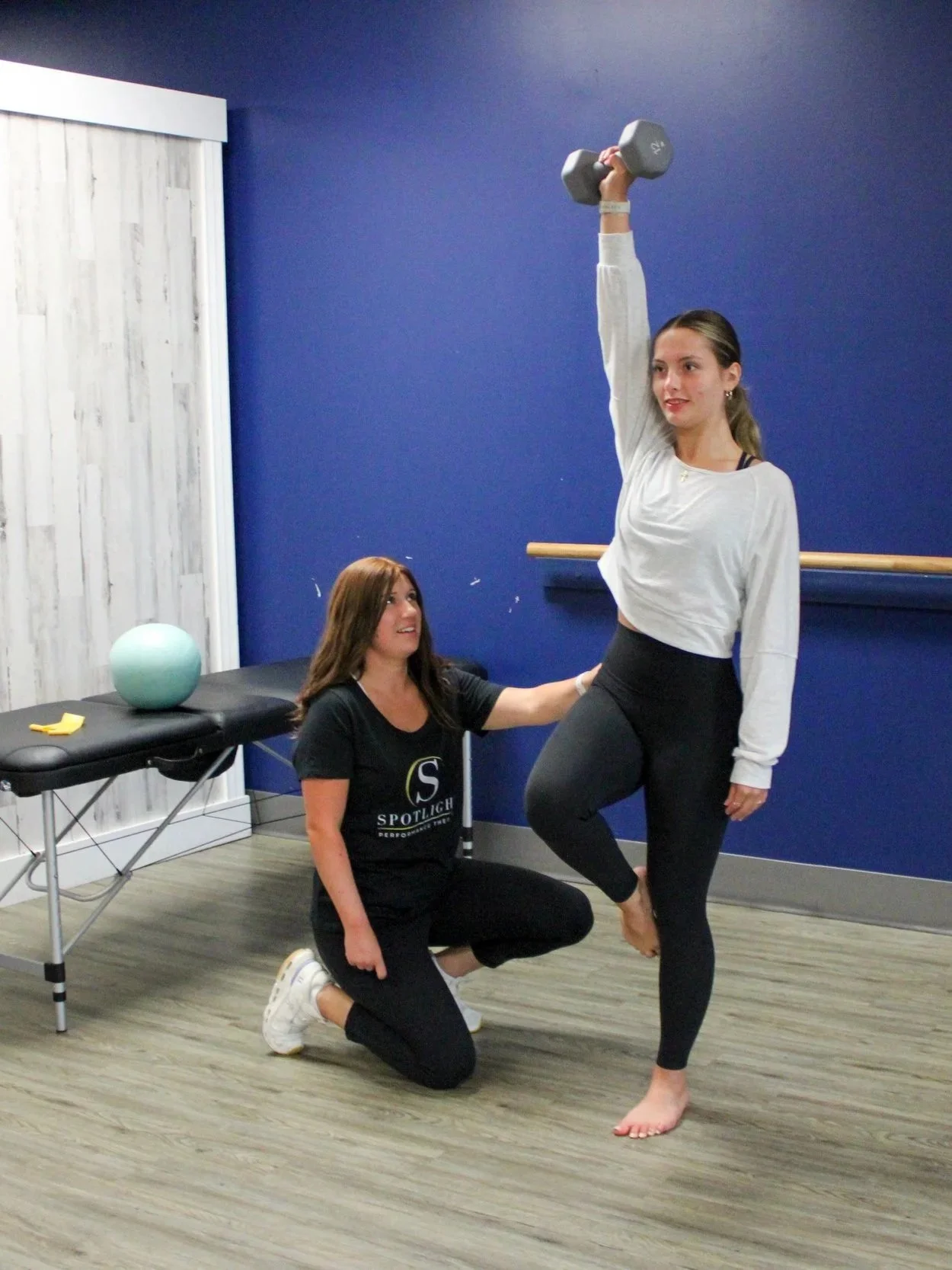 A woman in black leggings and a white shirt is practicing a yoga pose with a dumbbell raised in the air, assisted by a woman in black kneeling on the floor. The background includes a blue wall, a ballet barre, and a massage table with a therapy ball on top.