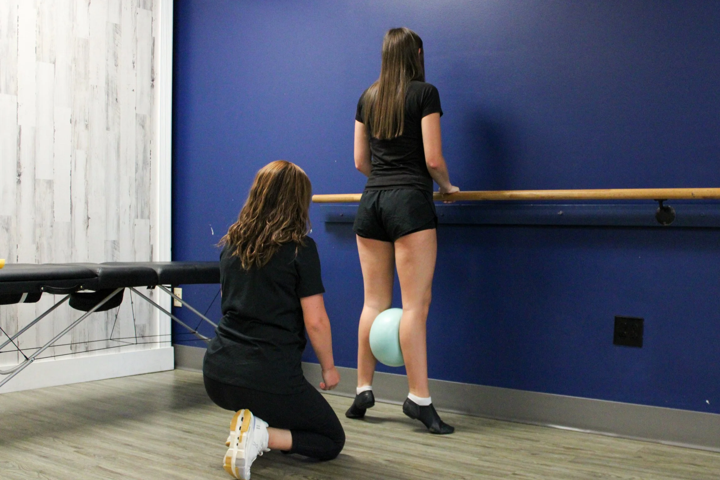 A woman kneeling on a wooden floor, with a person standing in front of a blue wall, holding a ballet barre. The woman appears to be instructing or observing as the person in front is preparing for ballet practice, with a ballet ball under their foot.