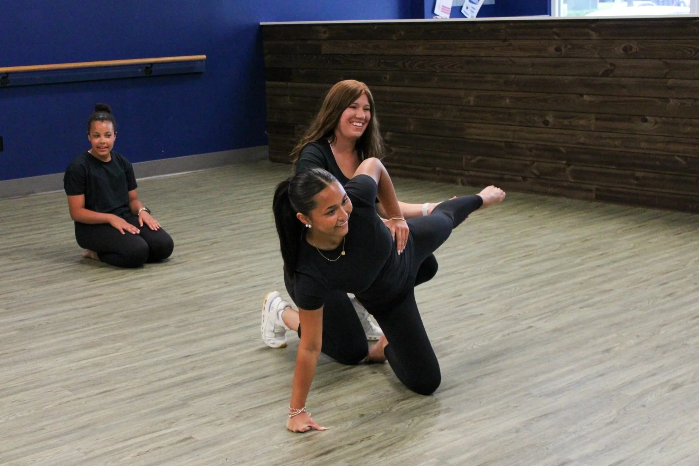 Two women practicing acro yoga, with the woman in the front supporting the woman in the back in a plank position, while a young girl kneels on the floor watching.