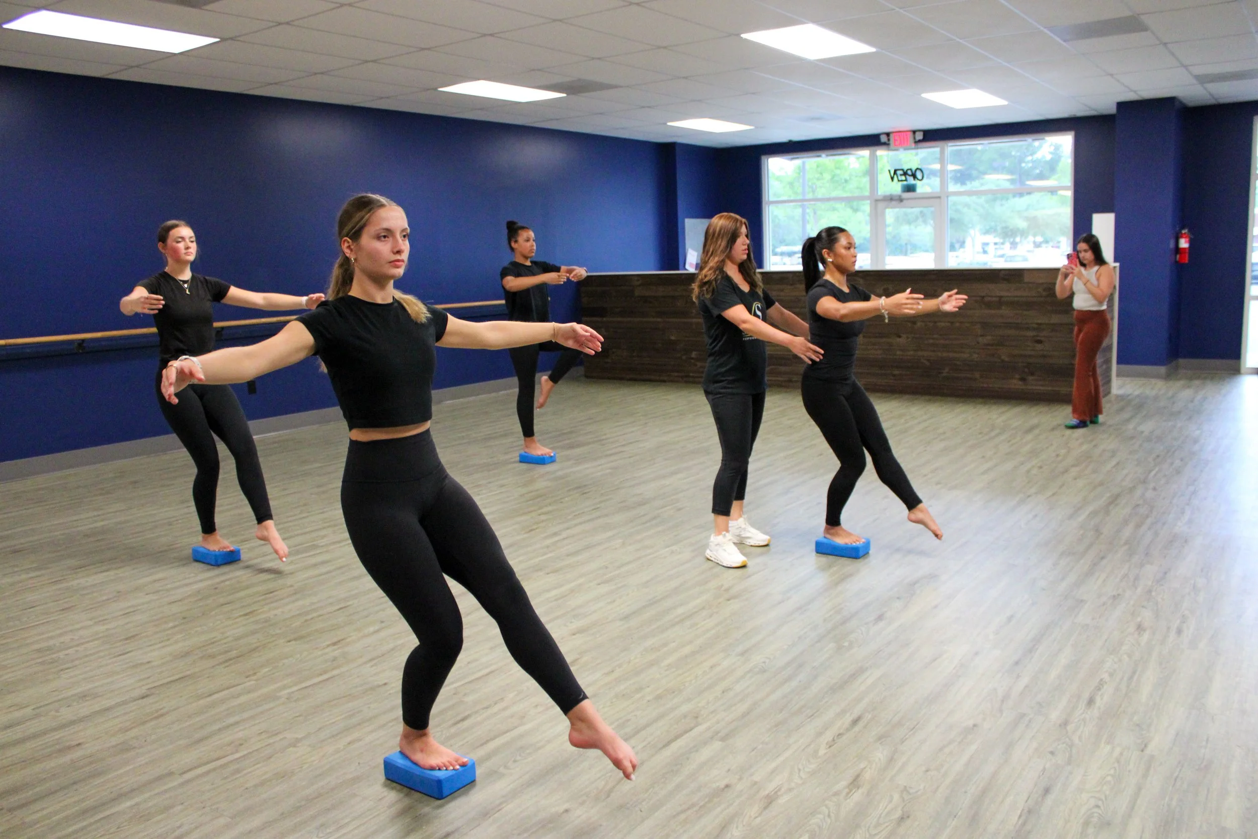 Five women practicing ballet in a studio, standing on blue balance blocks with arms extended, while a woman in the background takes a photo.