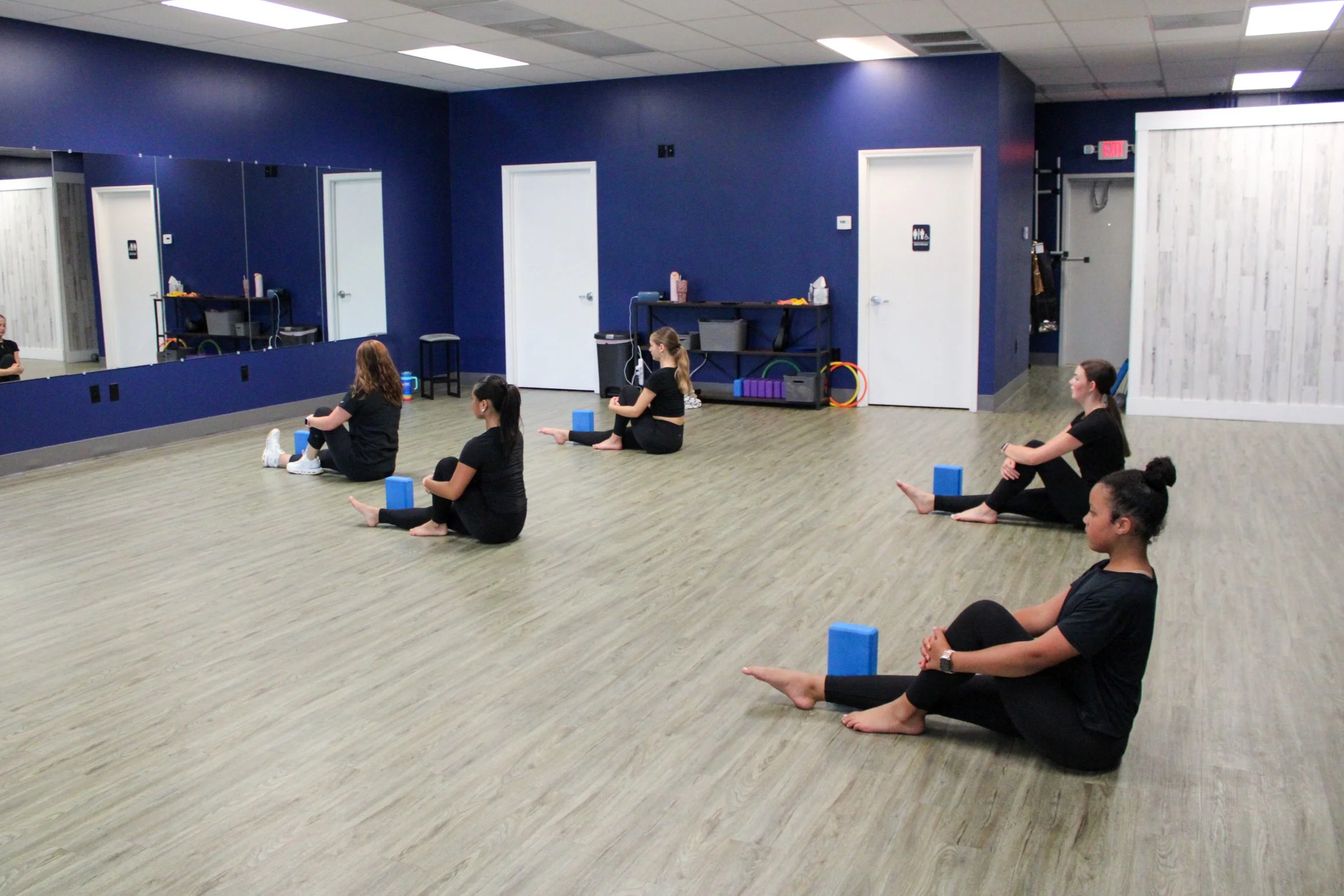 A group of women practicing yoga in a studio with blue walls, wooden floors, mirrors, and yoga props, seated with legs extended and arms resting on knees.