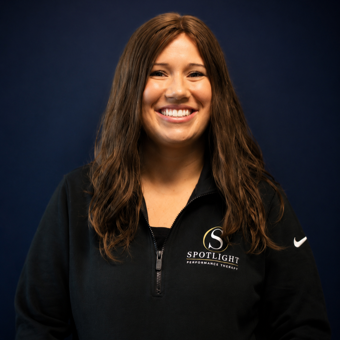 Smiling woman with long brown hair wearing a black jacket with the logo 'Spotlight Performance Therapy' and a Nike logo, standing against a dark blue background.