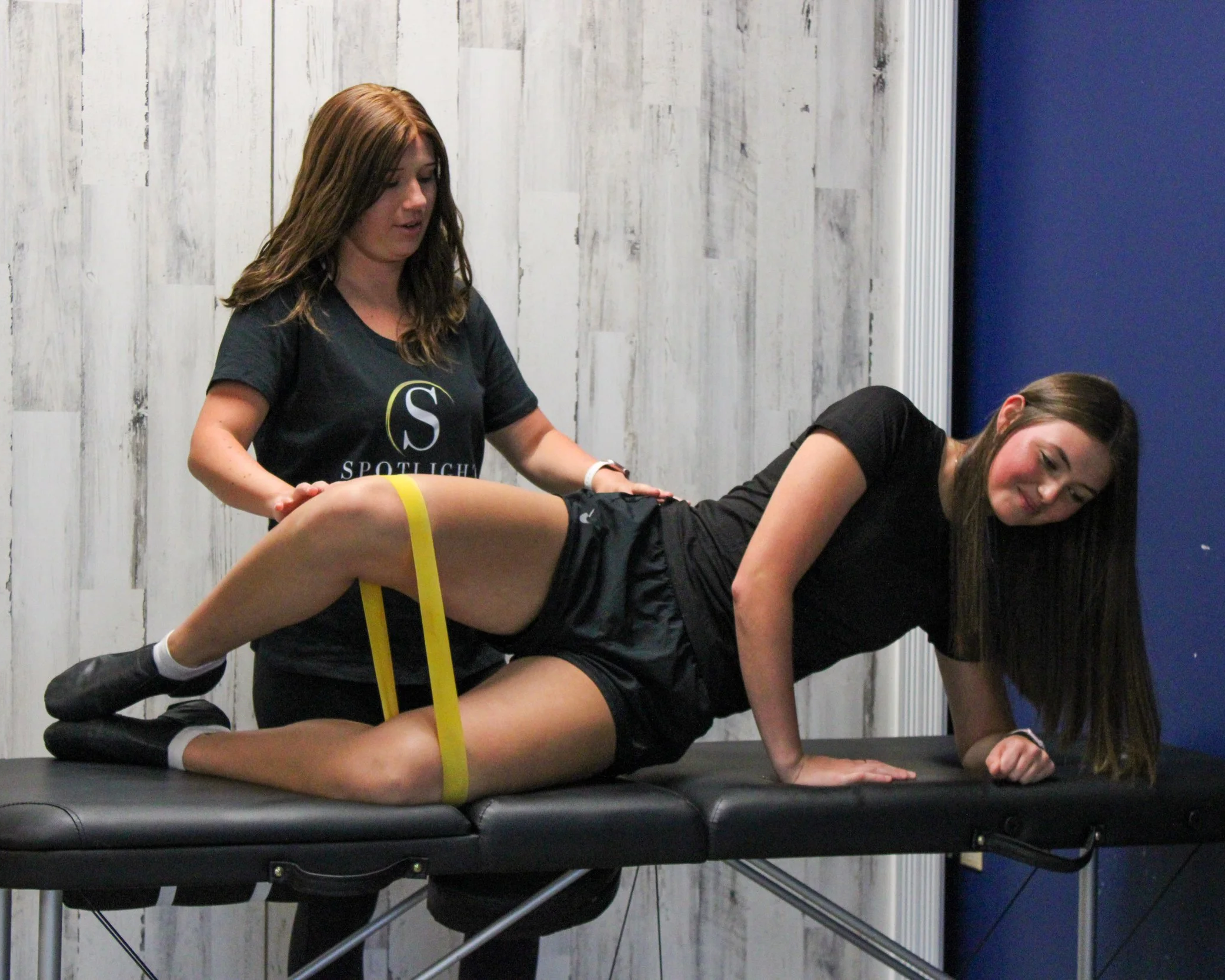 Two women in workout clothes practicing yoga or physical therapy on a black padded table in a room with white wooden panel walls and a blue wall. One woman is assisting the other, who is in a plank position with a yellow resistance band around her thighs.