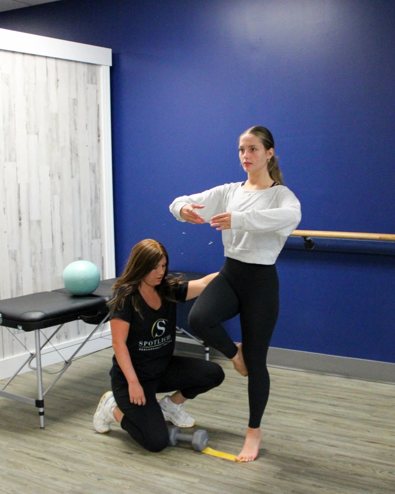 Two women in a fitness or physical therapy setting, one standing on one leg with a yellow tape on her foot, the other kneeling beside her, assisting with her balance. There is a small table with a teal exercise ball and a dumbbell.