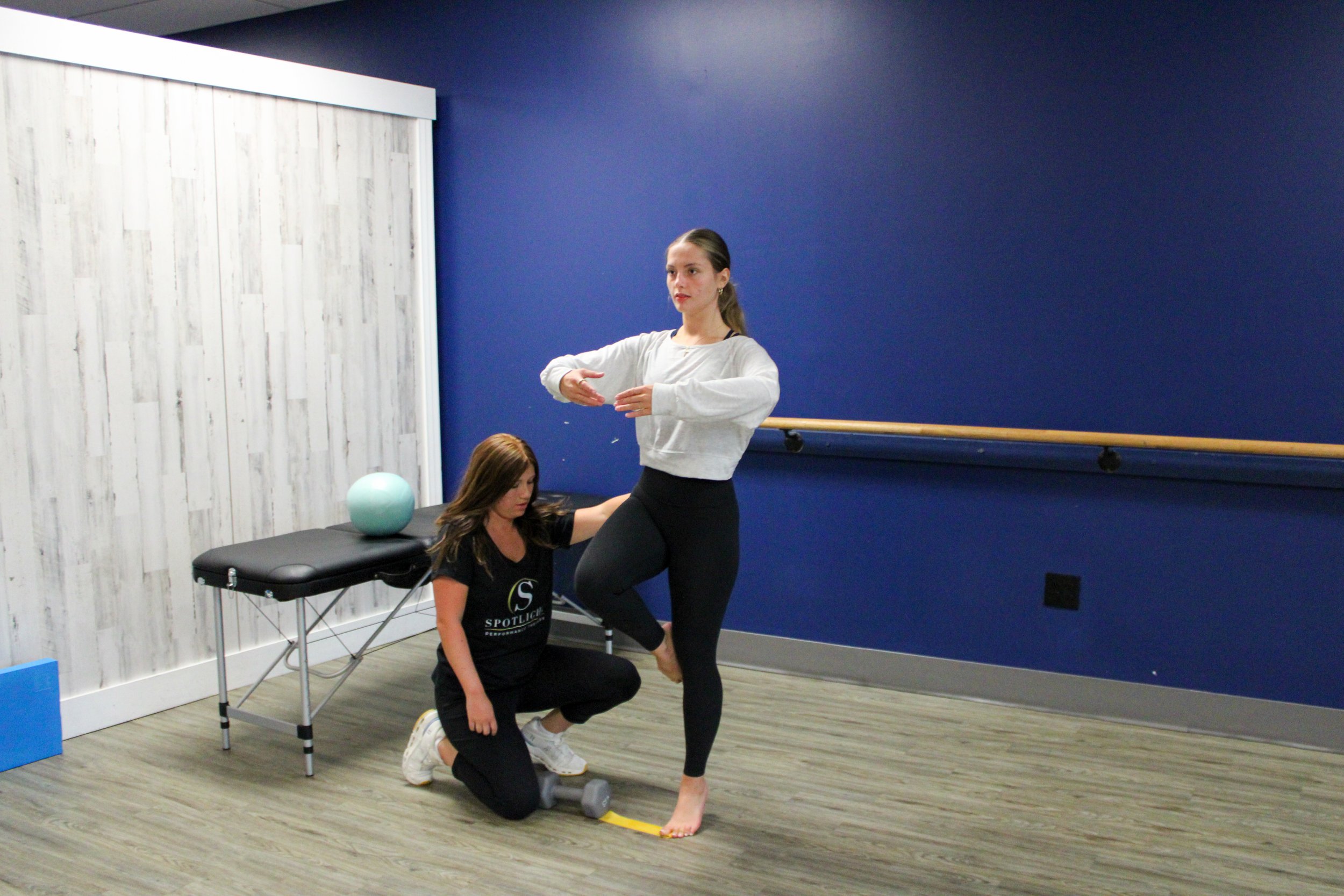 One woman is assisting another woman with a fitness or physical therapy exercise in a room with a blue wall and a wooden dance barre. The woman standing is holding her foot and has her arms crossed, while the woman kneeling is holding a dumbbell.
