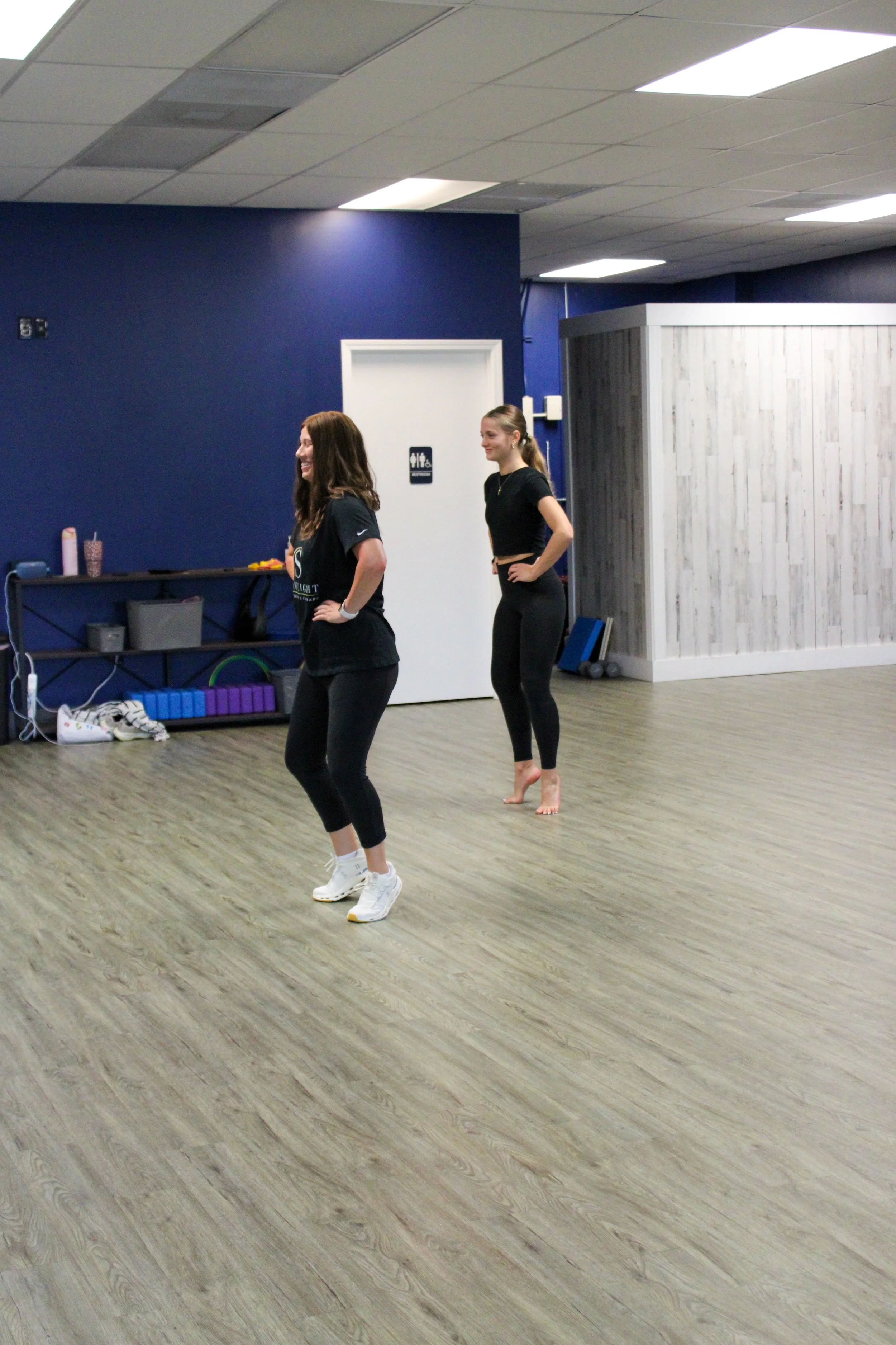 Two women participate in a fitness class in a room with dark blue walls and wood flooring, smiling and engaging in exercise.