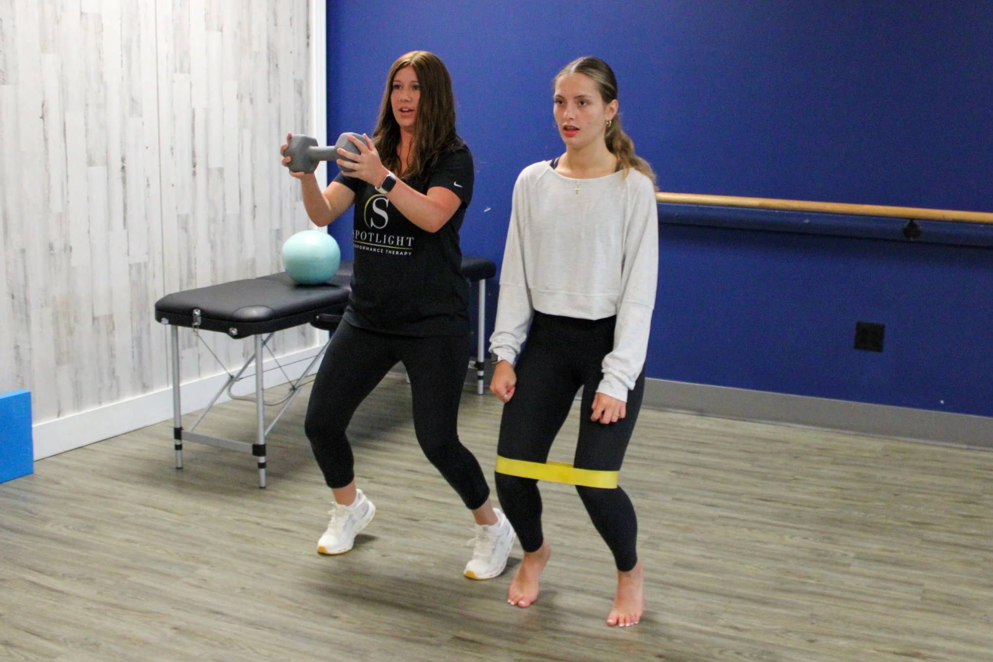 Two young women are in a fitness studio, one doing an exercise with a small dumbbell, and the other standing beside her. The woman with the dumbbell has long brown hair and is wearing a black T-shirt and black leggings. The woman beside her has light brown or blonde hair tied back, is wearing a white sweatshirt and black leggings, and is standing with her hands relaxed at her sides. The room has a wooden floor, a blue wall, a ballet barre, and a treatment table with a blue exercise ball on it.
