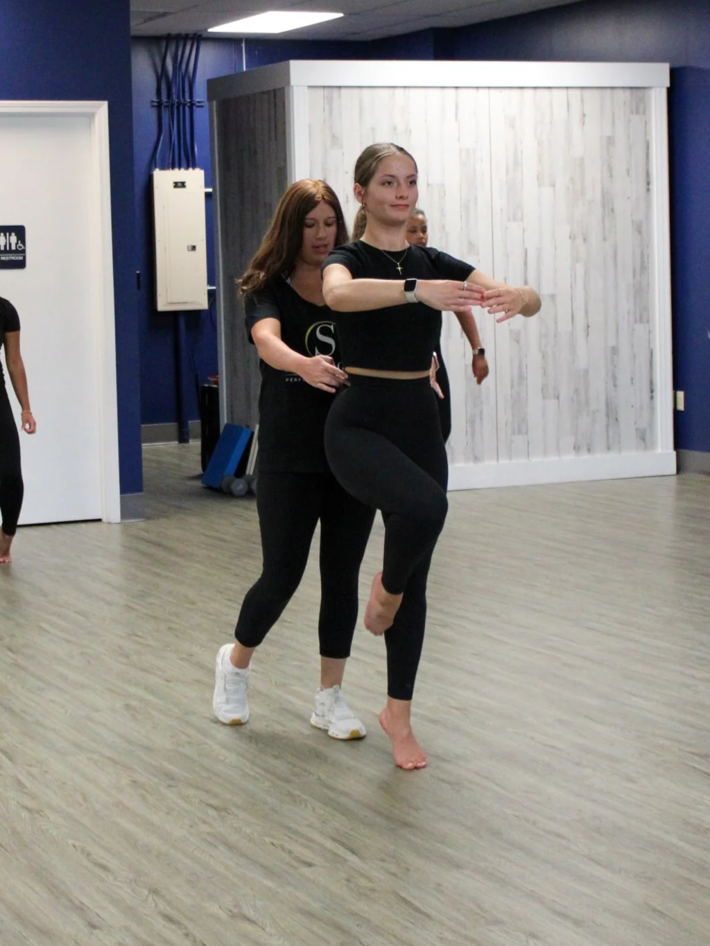 Two women practicing a dance move together in a studio. The woman in front is barefoot, balancing on one leg with arms extended, while the woman behind is supporting her. Both are wearing black athletic outfits.