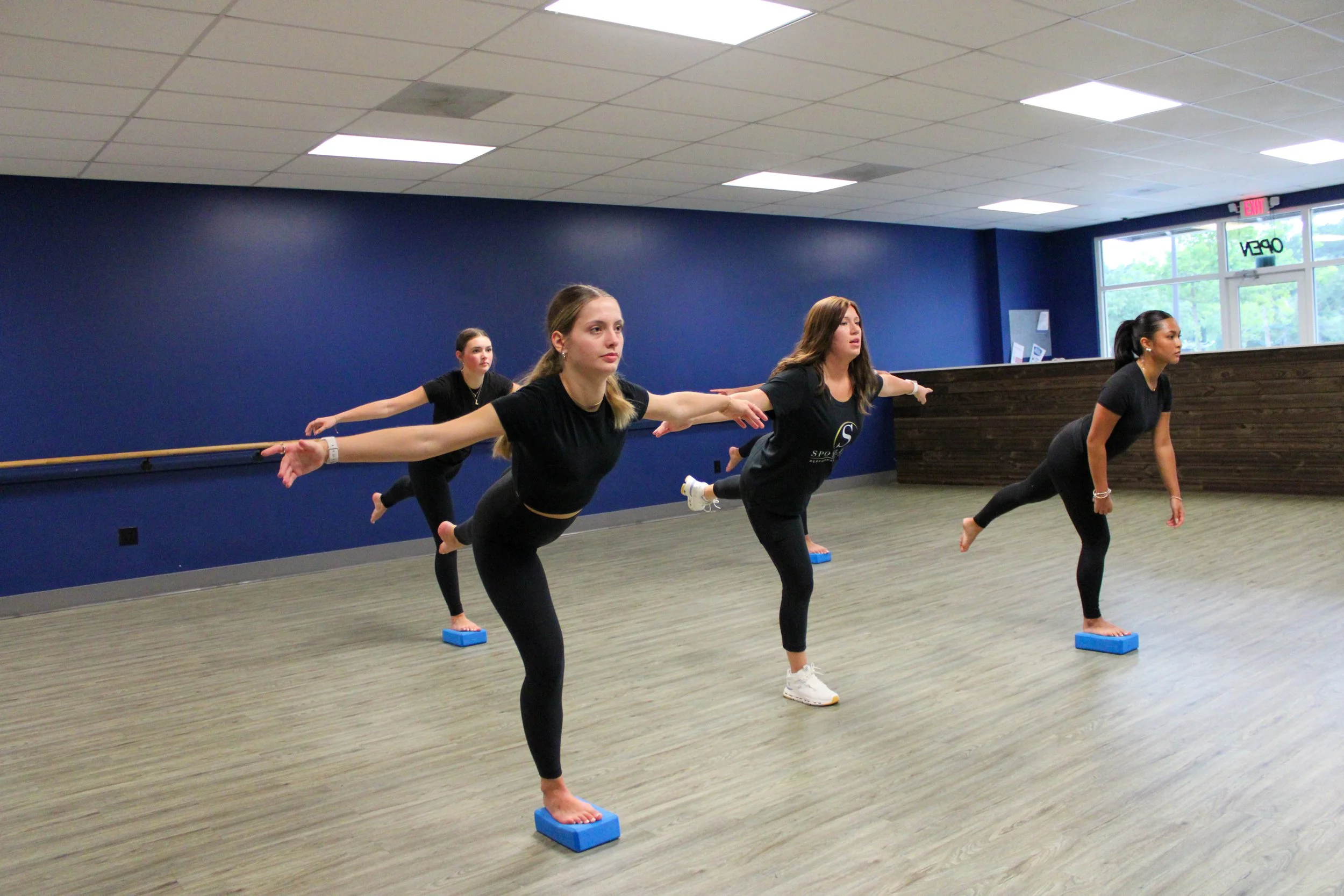 Four women practicing yoga in a dance studio, balancing on one leg with arms extended, using blocks for support, with mirrors and a blue wall in the background.