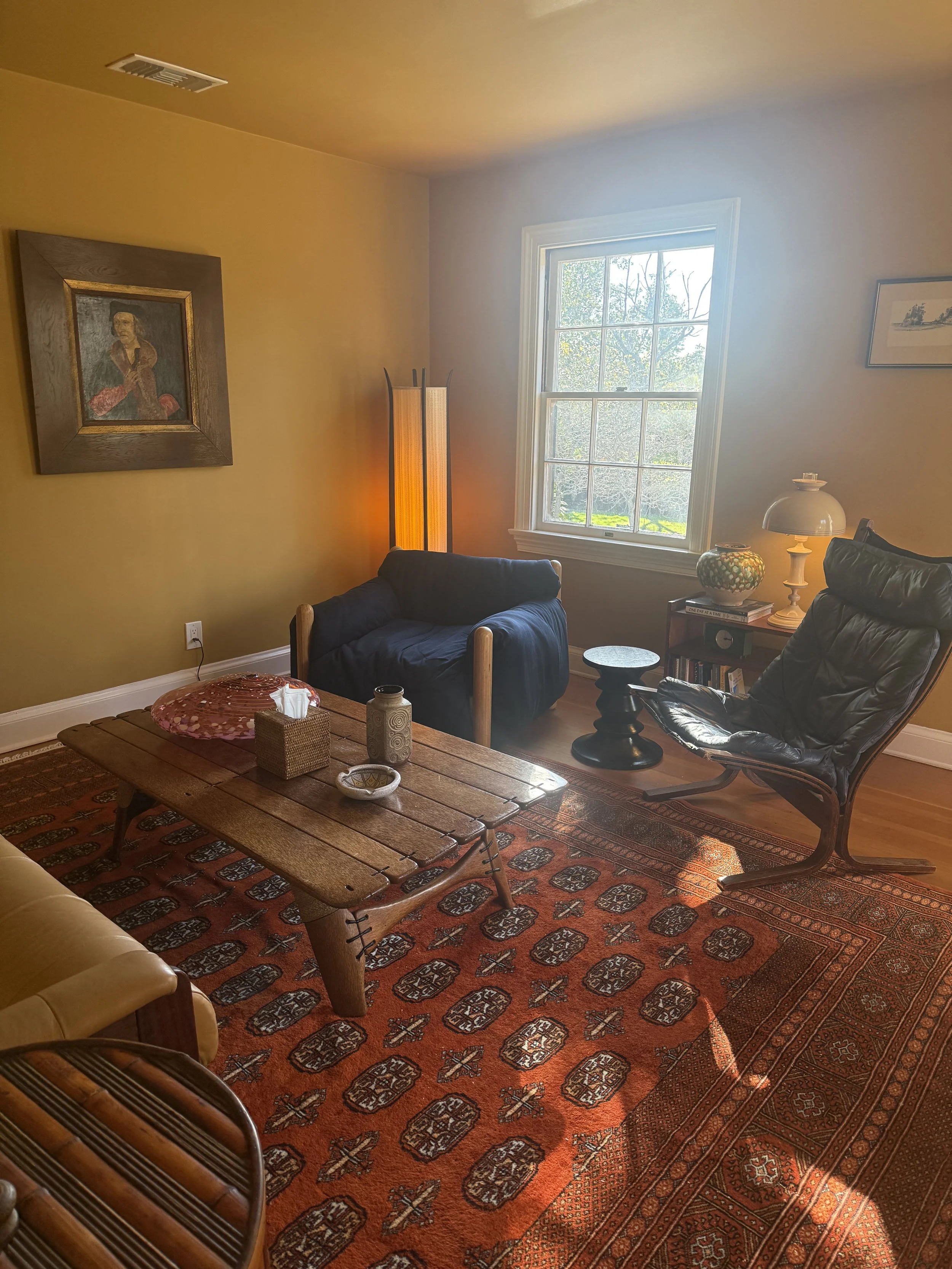 Therapy office with yellow and white walls, a window with trees outside, a wooden coffee table, a blue armchair, a small side table with a lamp and decorative item, a tall floor lamp, framed paintings, and an area rug.