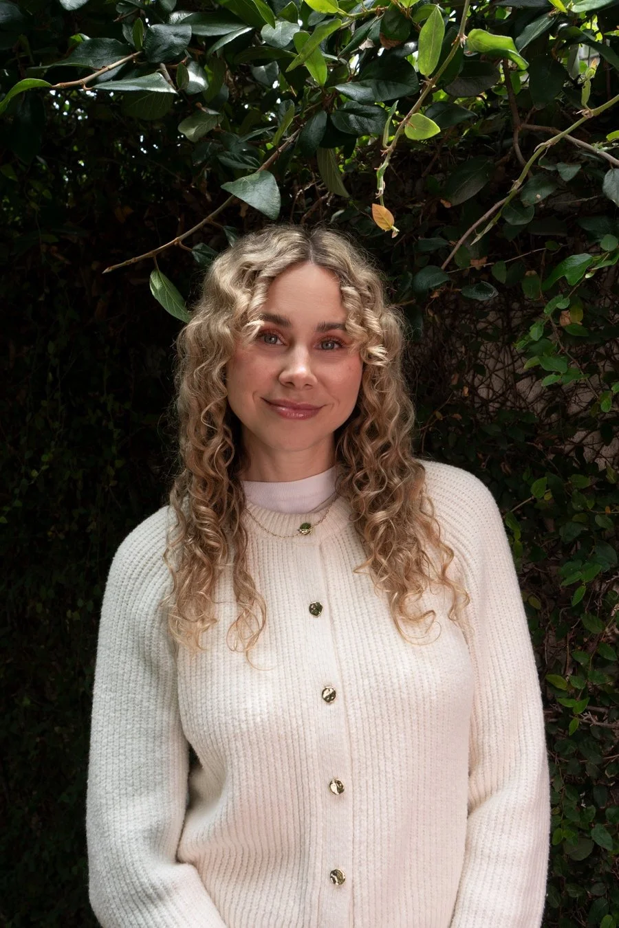 A woman with blonde curly hair and light skin, smiling, standing outdoors in front of green leafy bushes, wearing a cream-colored cardigan with gold buttons.