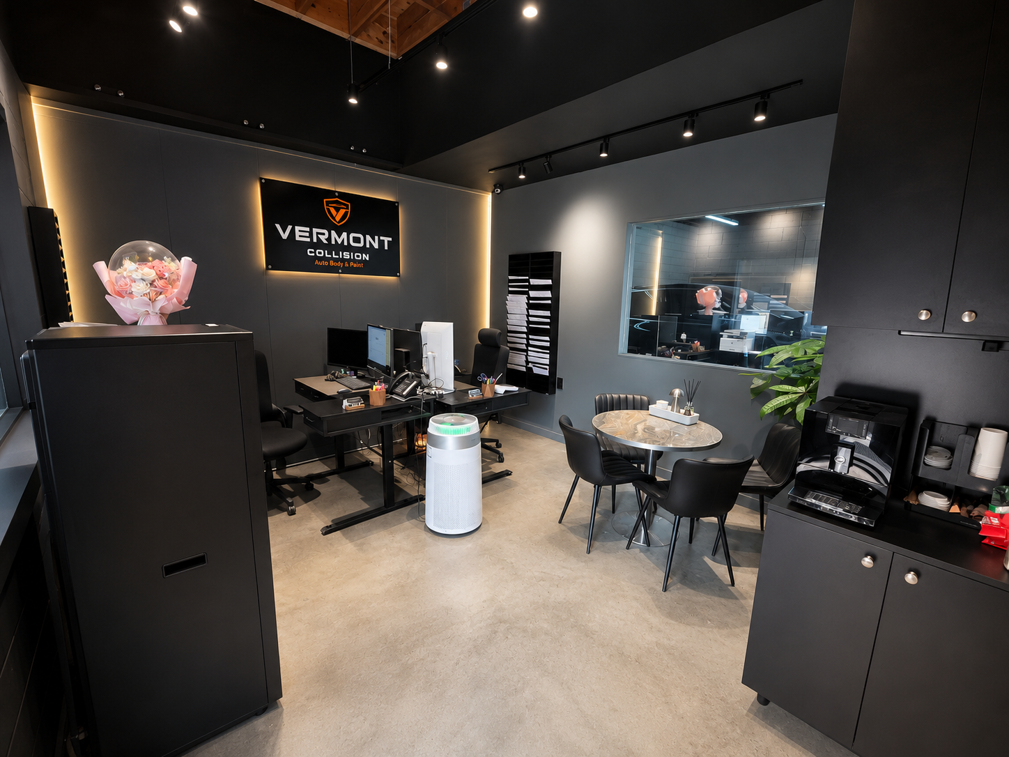Modern office reception area with a desk, computers, seating, and a large window looking into vehicle workshop, with a sign that reads 'Vermont Collision Auto Body & Paint'