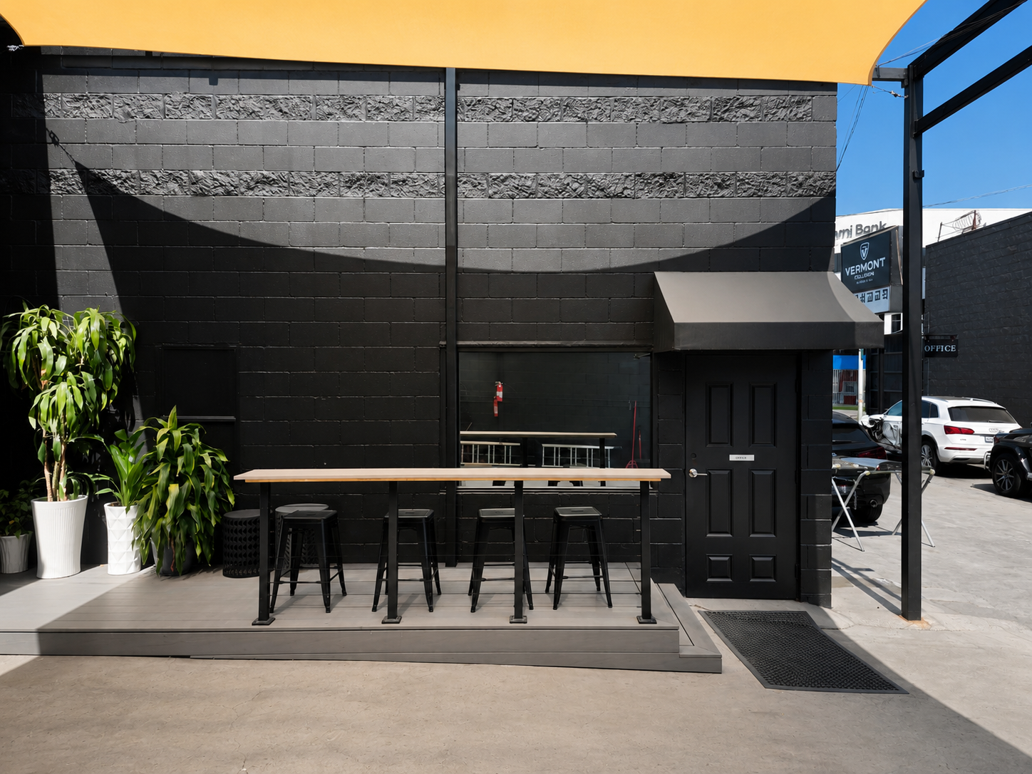Modern black brick building exterior with a small wooden deck, black stools, potted plants, and a windowed door with a black awning.