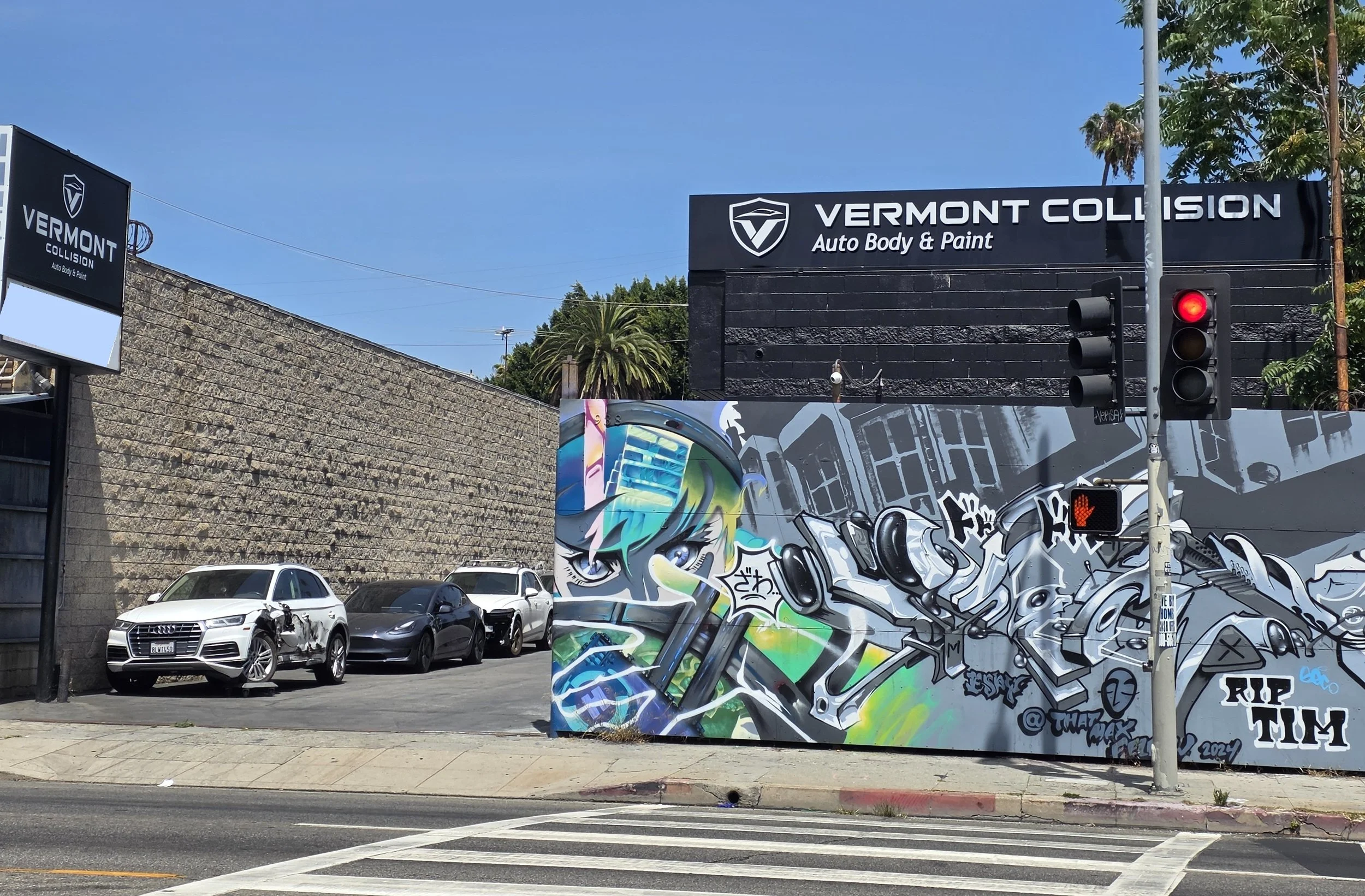 A streetcorner with a black building for Vermont Collision Auto Body & Paint, a traffic light with red light on, graffiti mural on a wall, multiple parked cars, and clear blue sky.
