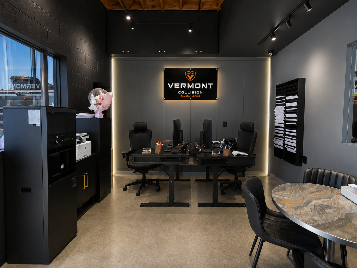 Modern reception area at Vermont Collision auto body shop with two black desks, office chairs, a sign on the wall, and a table with black chairs.