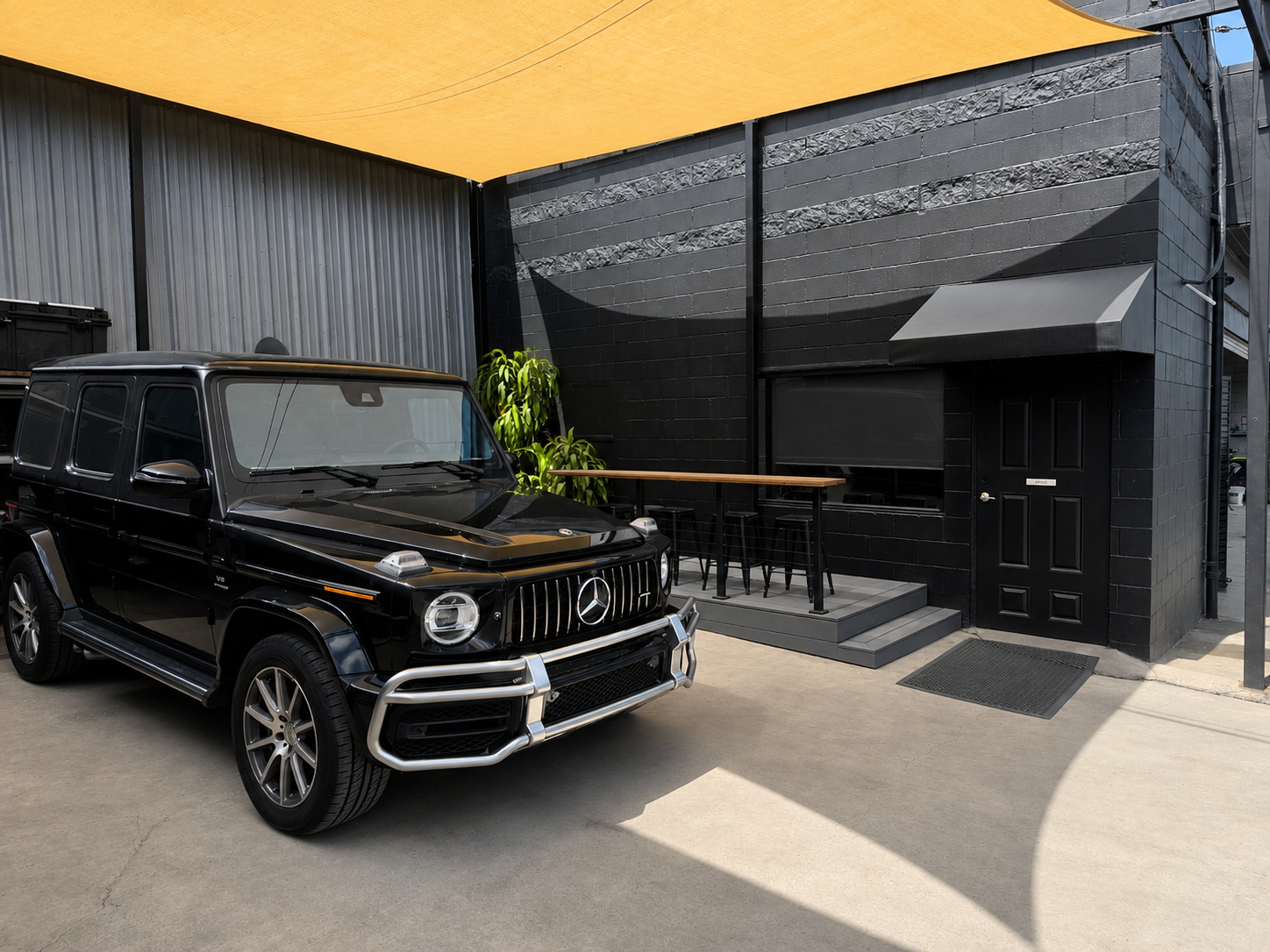 Black Mercedes-Benz G-Class SUV parked outside a black building with a small patio area featuring a tall plant, a high table, and stools, under a yellow shade sail.