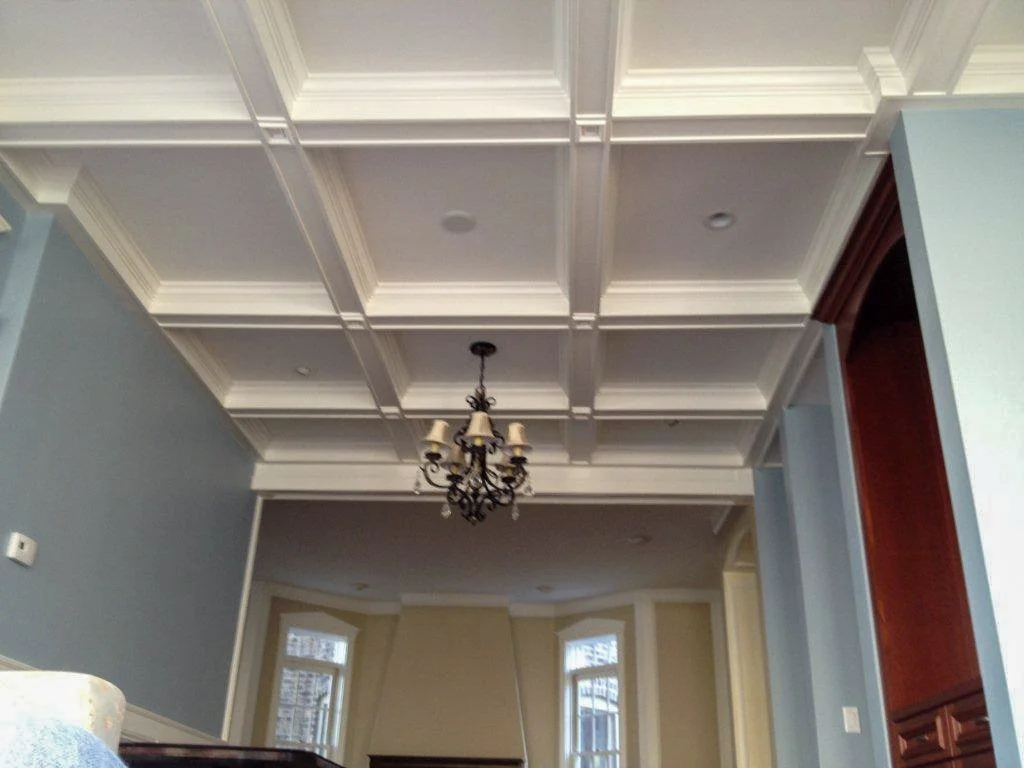 Image of a ceiling with decorative beams and a chandelier in a living room with large windows.