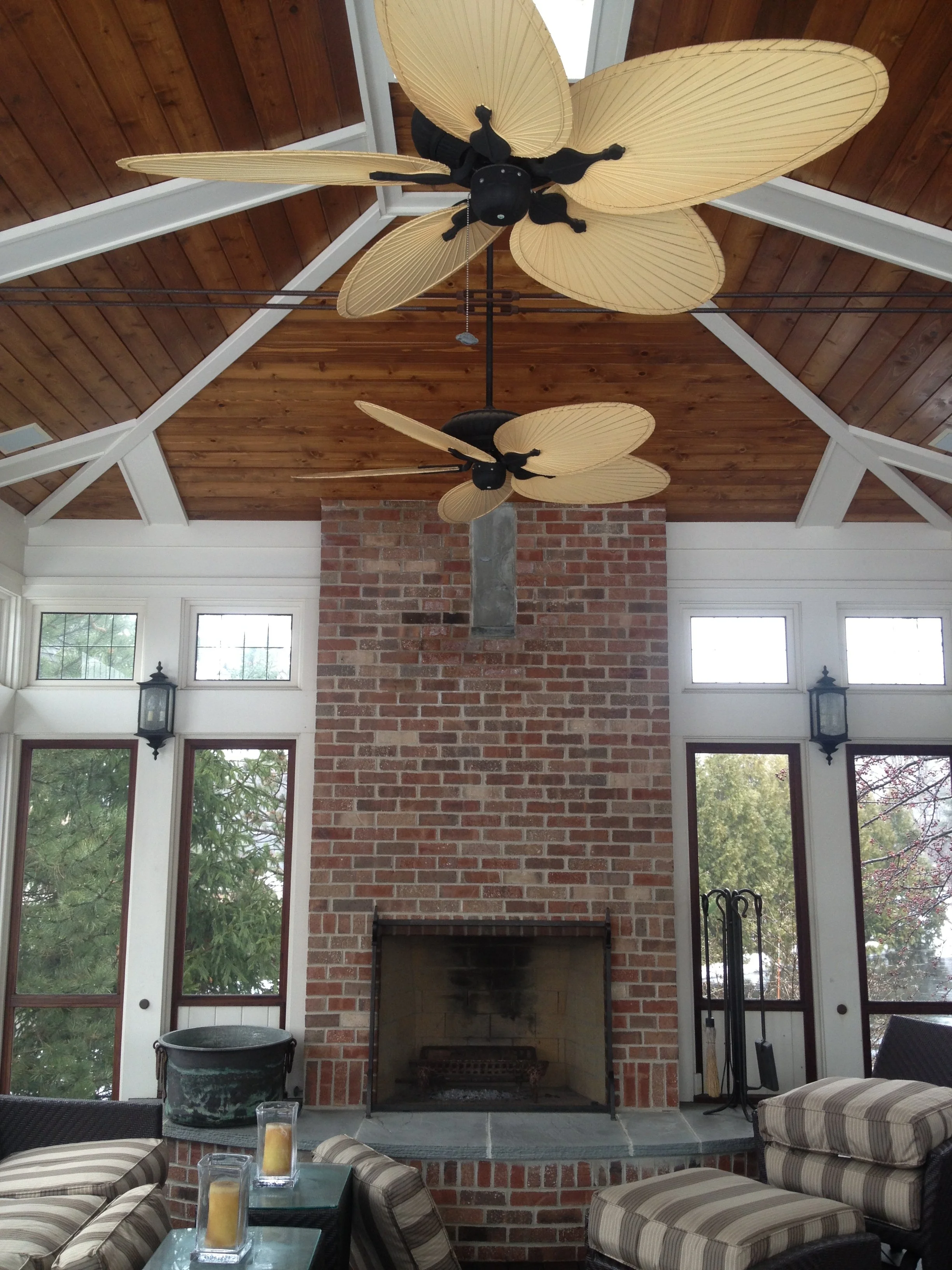 Indoor space with a tall brick fireplace, ceiling fans with leaf-shaped blades, large windows, and striped cushioned chairs arranged around a glass table with candles inside glass holders.