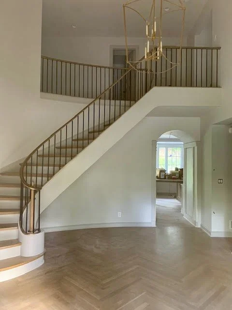 Empty living room with a staircase and gold railing leading to an upper level, chandelier hanging from the ceiling, and a window in the background.