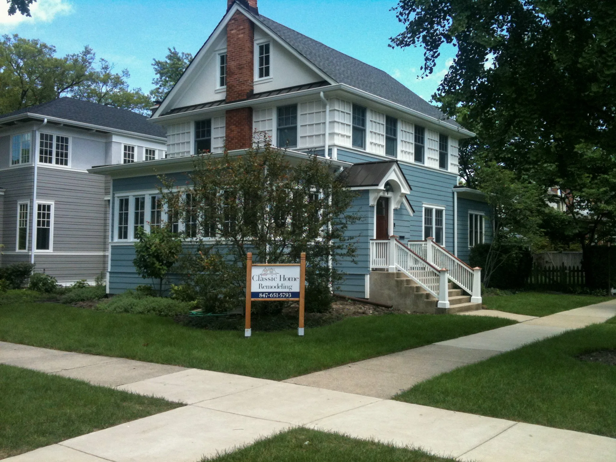 A blue and white two-story house with a brick chimney, surrounded by trees and a well-maintained lawn. There's a sidewalk in front and a sign in the yard advertising home remodeling services.