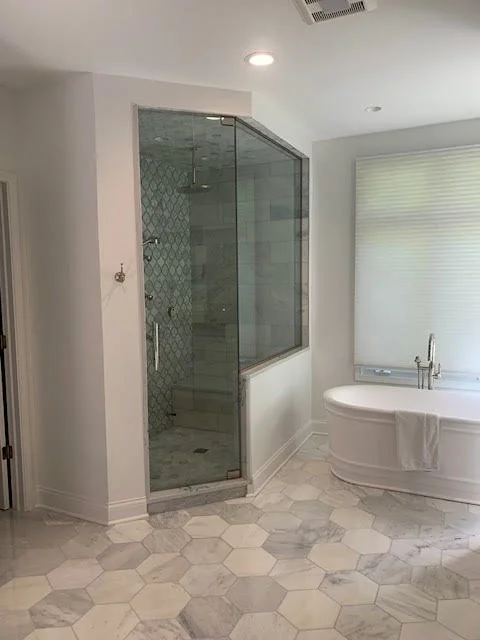 Bathroom with a glass-enclosed shower, a white bathtub with a towel, marble tile flooring, and a window with blinds.
