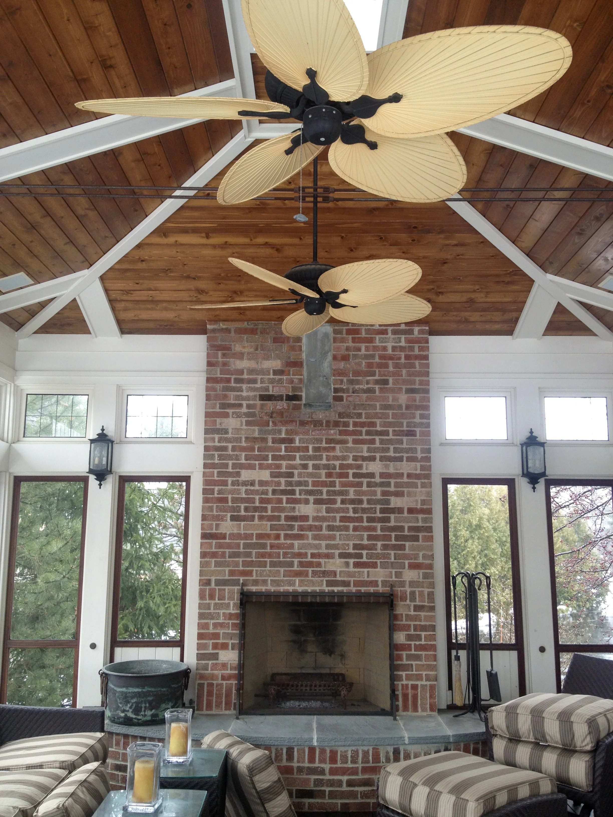 Living room with brick fireplace, four large windows, ceiling fans with beige leaf-shaped blades, wood-paneled ceiling, and framed by white trim. Furniture includes striped cushioned chairs and a glass coffee table with candles.