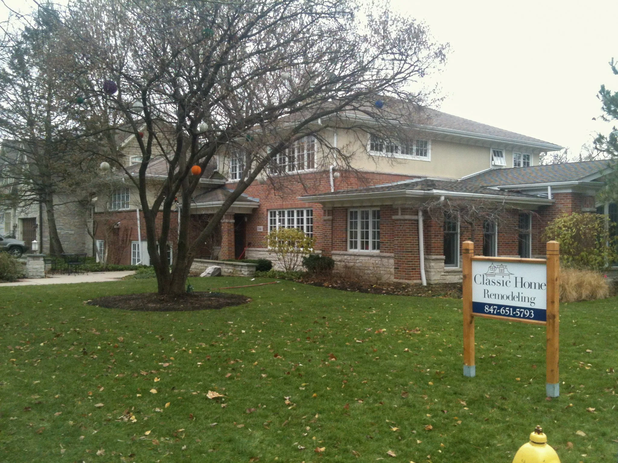 A residential house with a brick and beige exterior, multiple windows, and a landscaped front yard with grass and fallen autumn leaves. There is a sign that reads "Classic Home Remodeling" with a phone number, and a tree in the yard is decorated with