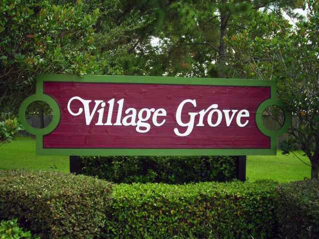 Sign that reads 'Village Grove' in white letters on a purple background with green border, surrounded by trees and bushes.