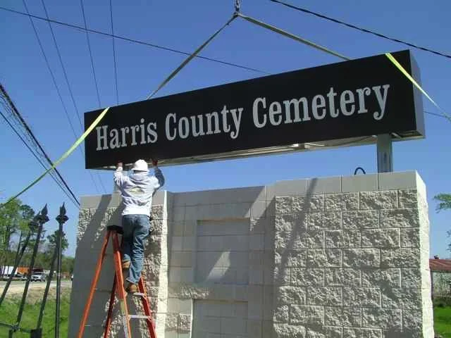 Person on a ladder installing or repairing a large black sign that reads "Harris County Cemetery" under a clear blue sky.