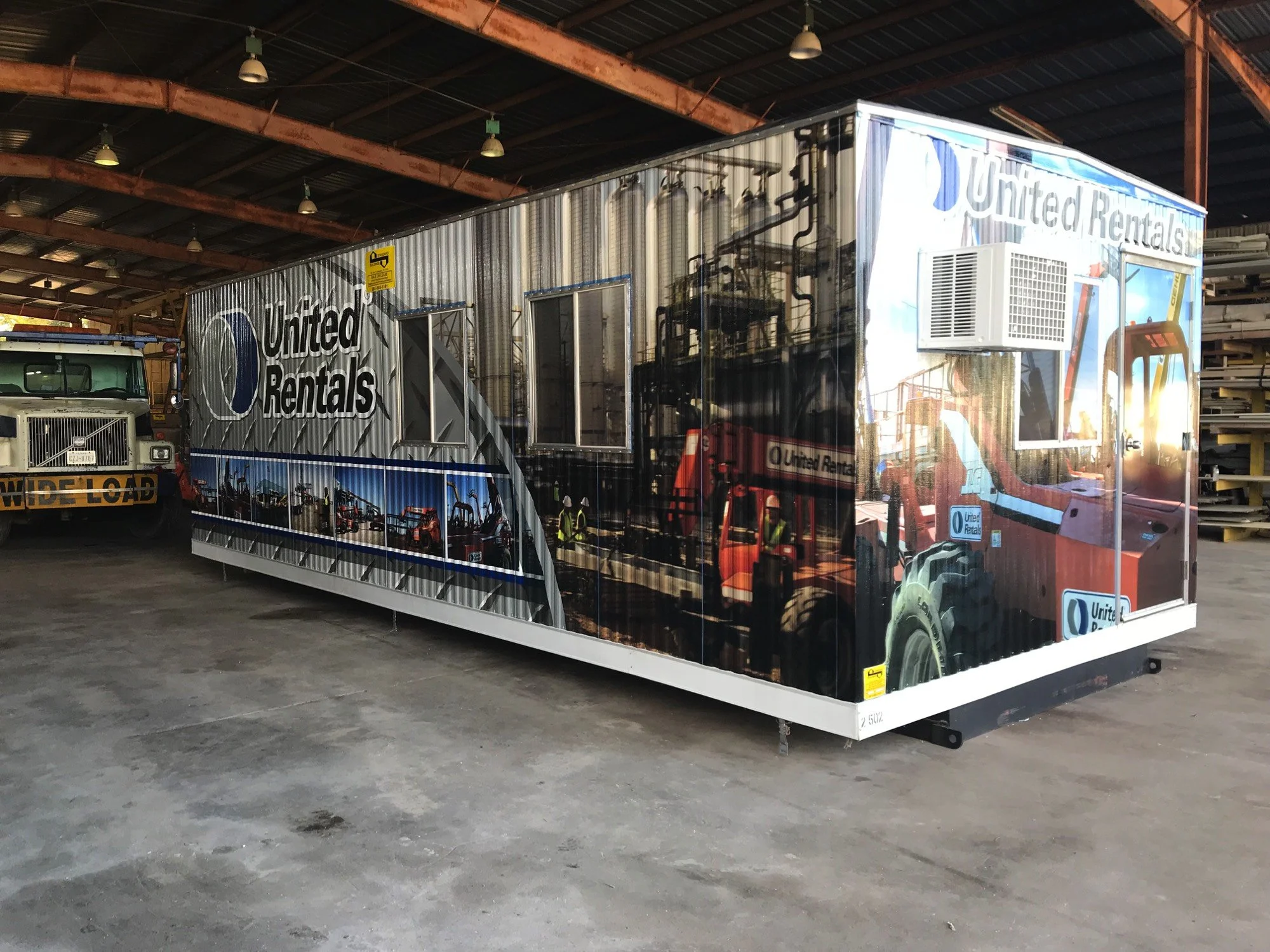 A large, reflective U-Haul truck inside a warehouse with a high ceiling and metal beams. Part of another truck is visible on the left, and wooden materials are stacked on the right side.