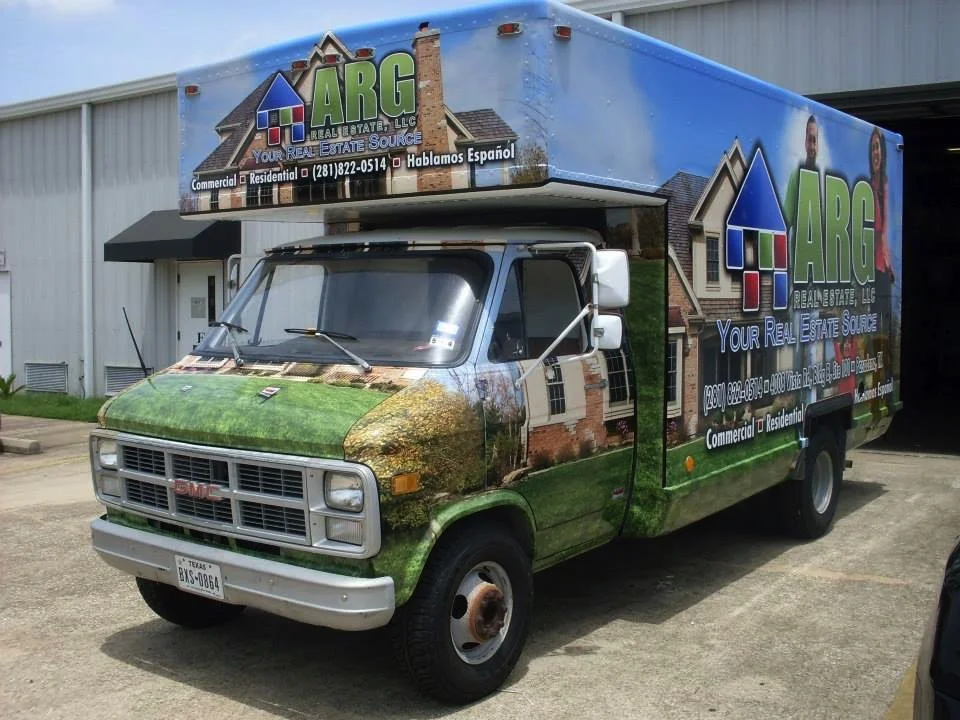 A GMC moving truck with advertising for ARG Real Estate, LLC, featuring images of houses and a grassy lawn, parked outside a building.