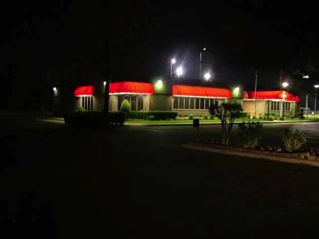 Nighttime view of a building with a red roof, illuminated by streetlights, with a parking lot and landscaped bushes in the foreground.