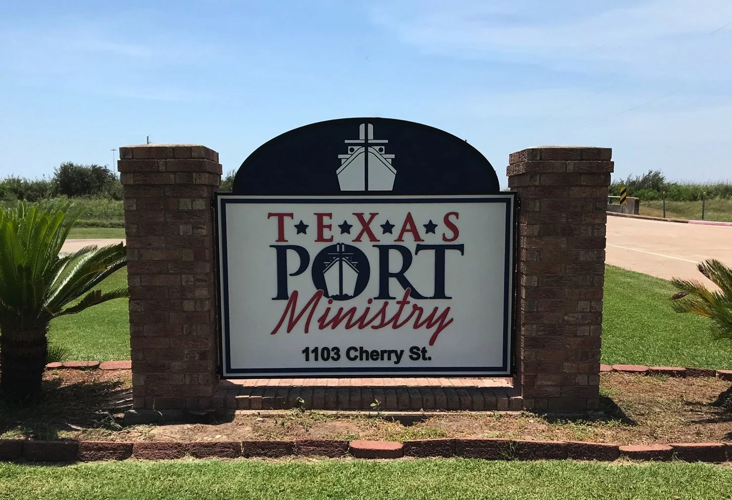 Sign reading 'Texas Port Ministry' with an illustration of a ship's bow at the top, located at 1103 Cherry St., framed by brick columns in a grassy area.