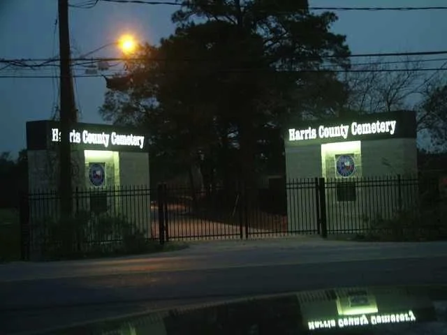 Entrance gates to Harris County Cemetery with illuminated signboards on each side, surrounded by trees and a fence, taken during dusk or dawn.