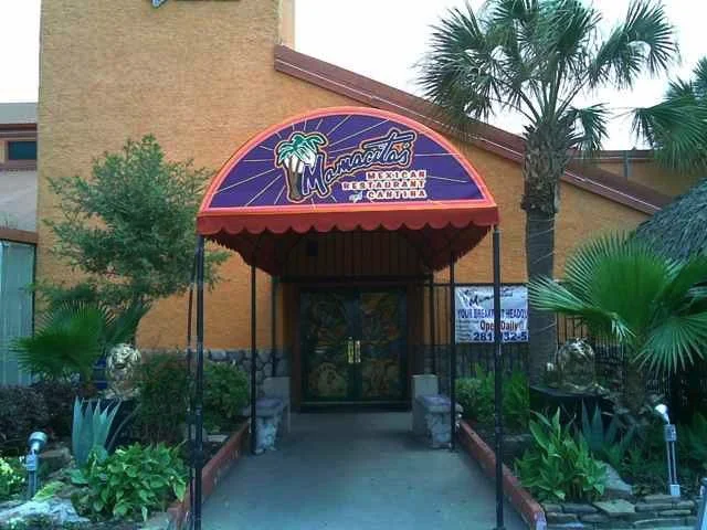 Entrance to Mamacitas Mexican Restaurant with an orange awning, tropical plants, palm trees, and a sign indicating the restaurant is open daily.