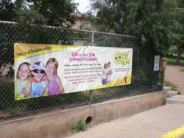 A banner on a chain-link fence advertising summer programs for YMCA of Boulder Valley, featuring images of children, with trees and a path in the background.
