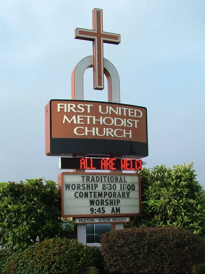 Sign for First United Methodist Church with a large cross on top, displaying worship service times and a message that all are welcome.