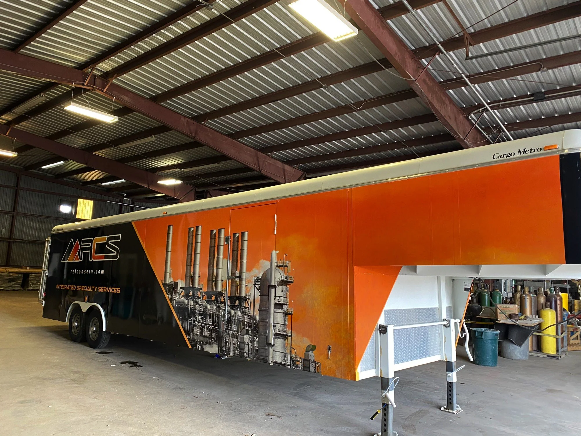 An orange trailer with industrial equipment graphic and the text 'Cargo Metro' parked inside a warehouse with a metal roof and fluorescent lighting.
