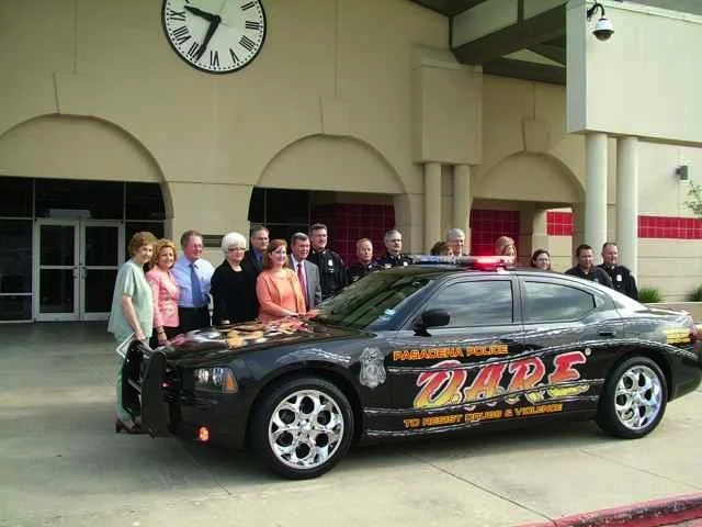 A group of people, including some in uniform, standing behind a black police car with a sunroof and police insignia, parked in front of a building with a clock showing 9:04.