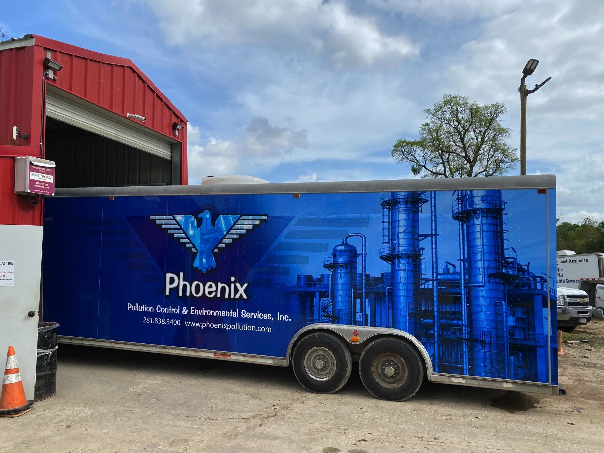 A blue truck with Phoenix Pollution Control & Environmental Services Inc. branding parked on a dirt lot. The logo features a stylized bird with outstretched wings. In the background, a red building, a tall light pole, a large tree, and some vehicles 